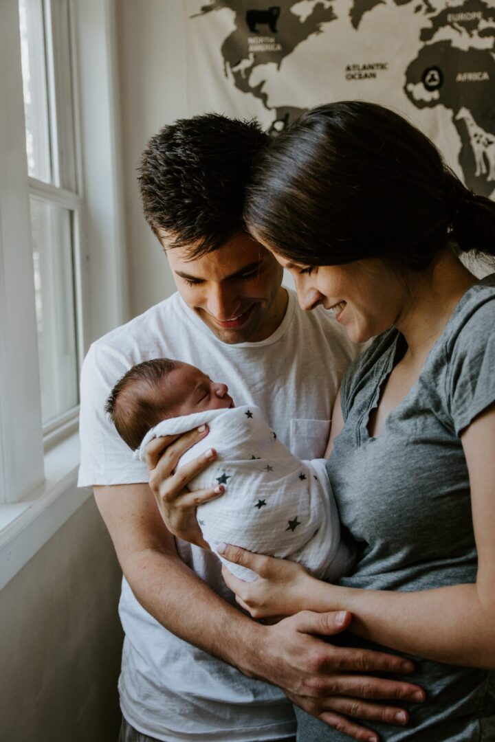 A man and woman holding a baby in front of a window.